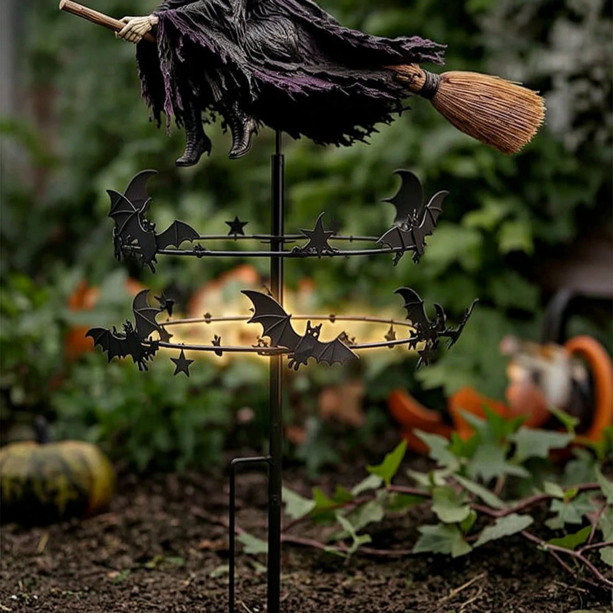 A black bat wind rotator garden decor featuring a dark fabric and a broomstick on a metal stand.