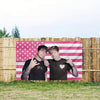 A pink American flag tapestry featuring stars and stripes displayed outdoors against a wooden fence.