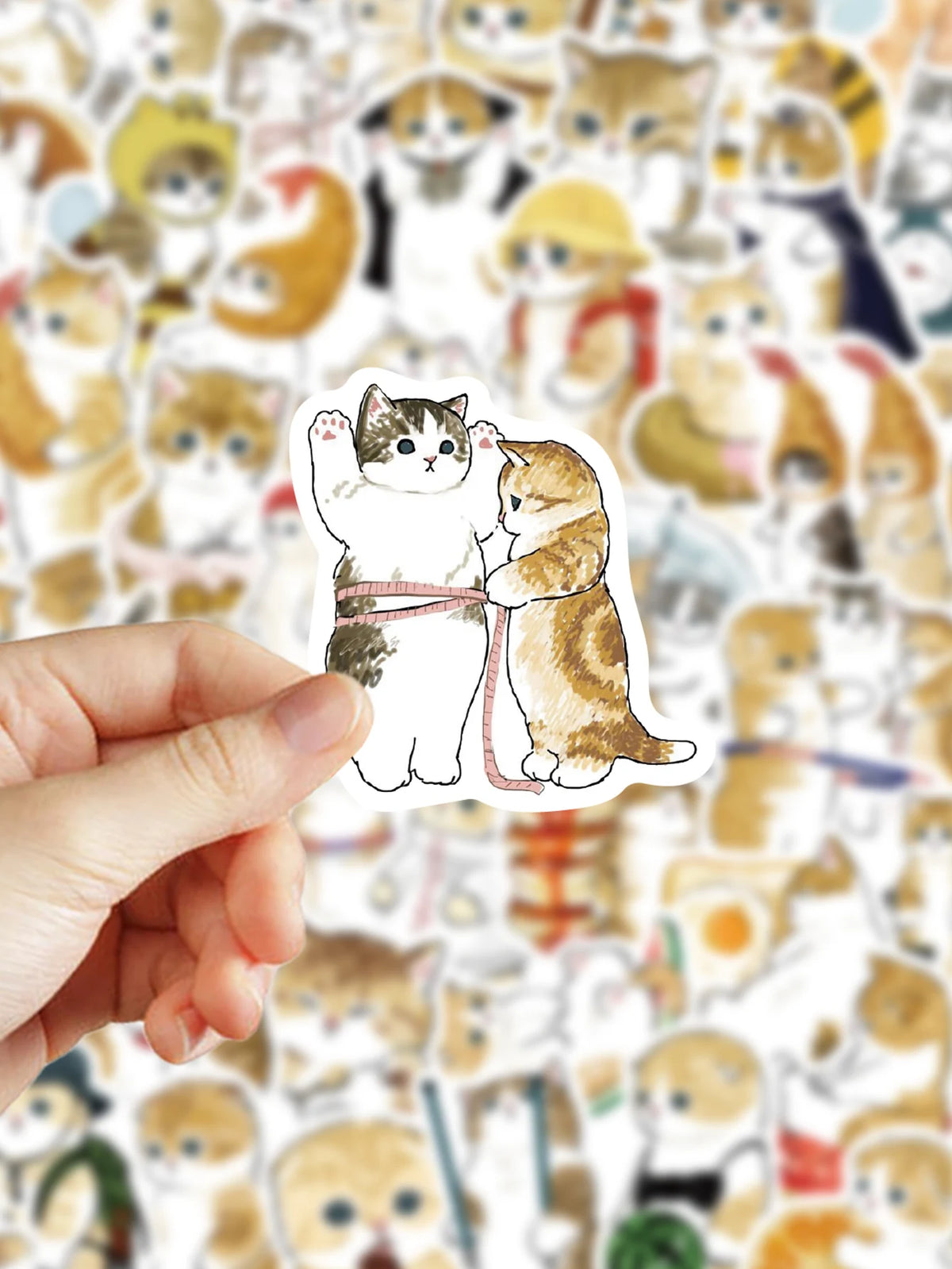 A hand holds a cat sticker featuring two kittens hugging against a background of many cute cats.