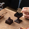 A hand holds a dark brown Chinoiserie Square Pixiu incense burner with smoke rising from it on a wooden surface.
