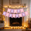 A pink and purple birthday garland banner strung across a fireplace mantel with warm string lights.