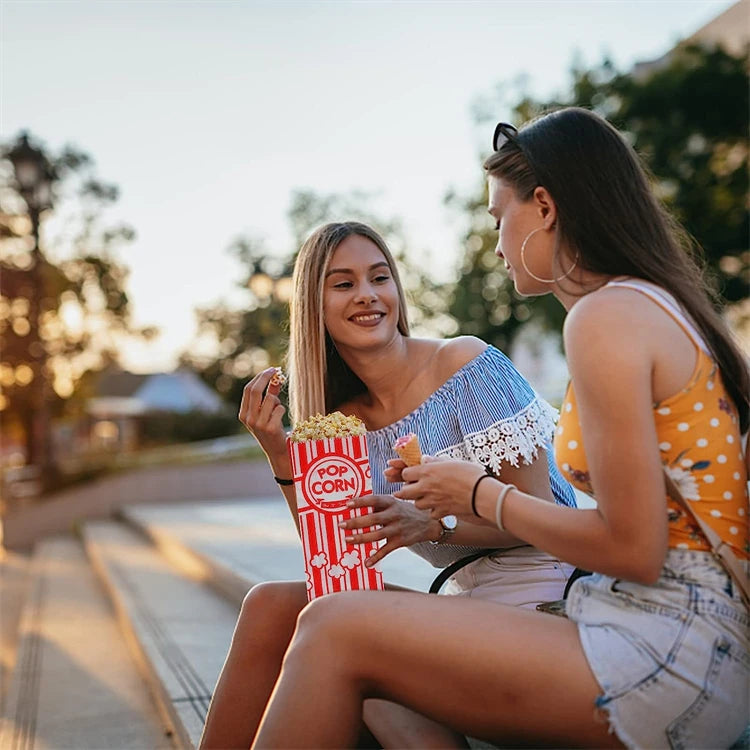 A young woman is holding a red and white popcorn bag while sitting outdoors during sunset.