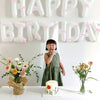 A child stands in front of a white balloon banner spelling out HAPPY BIRTHDAY with pink ribbons.