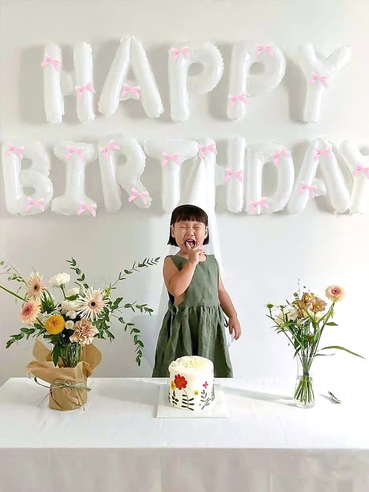 A child stands in front of a white balloon banner spelling out HAPPY BIRTHDAY with pink ribbons.
