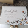 A white personalized wedding guest book with elegant script and floral watercolor design on a wooden surface.