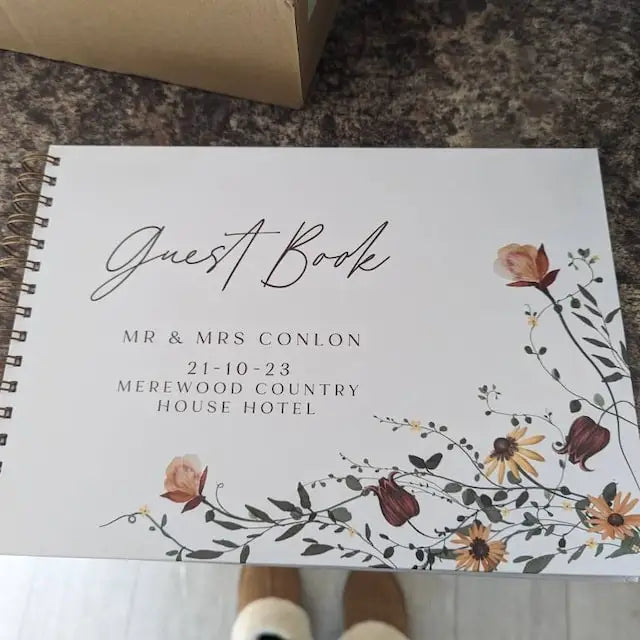 A white personalized wedding guest book with elegant script and floral watercolor design on a wooden surface.