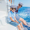 A smiling woman wearing sunglasses and a striped shirt is sitting on a boat railing with blue water in the background.
