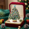 A rhinestone Christmas tree brooch pin displayed in a red velvet box with pine needles and gold accents.