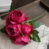 A bouquet of deep pink artificial silk peonies with green leaves resting on a beige linen fabric.