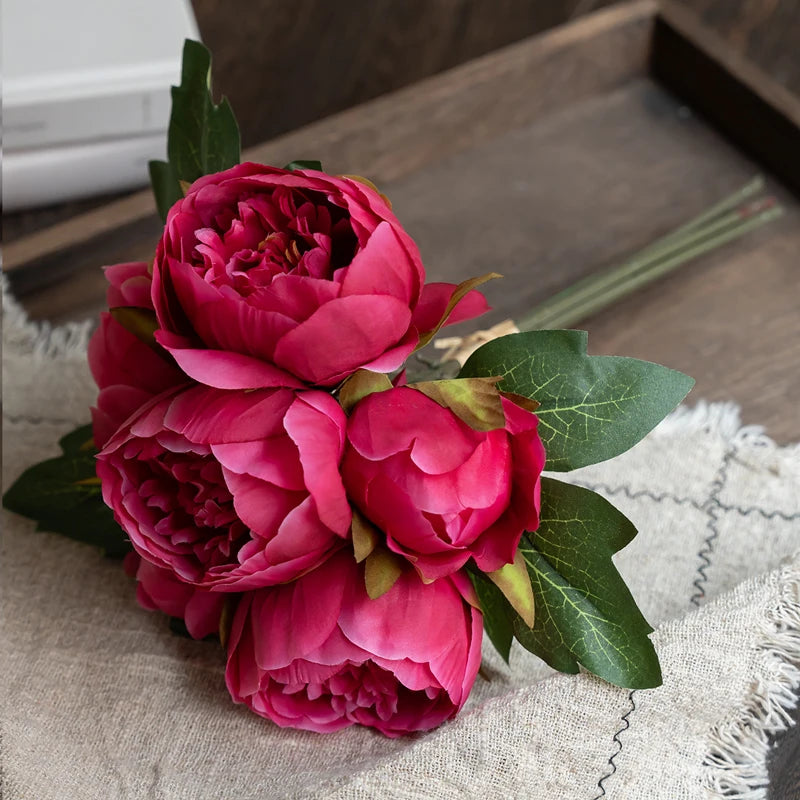 A bouquet of deep pink artificial silk peonies with green leaves resting on a beige linen fabric.