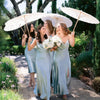 Three women in teal dresses hold white oil paper umbrellas while walking through a garden pathway.