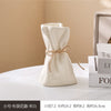 A beige ceramic money bag shaped vase is shown on a white surface next to a stack of books.