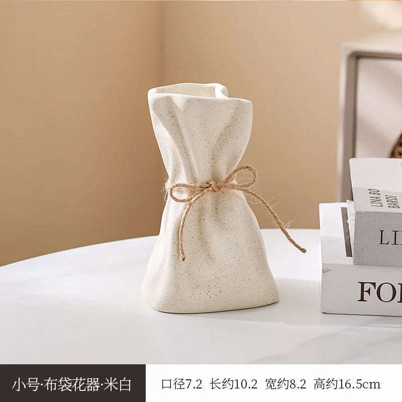 A beige ceramic money bag shaped vase is shown on a white surface next to a stack of books.