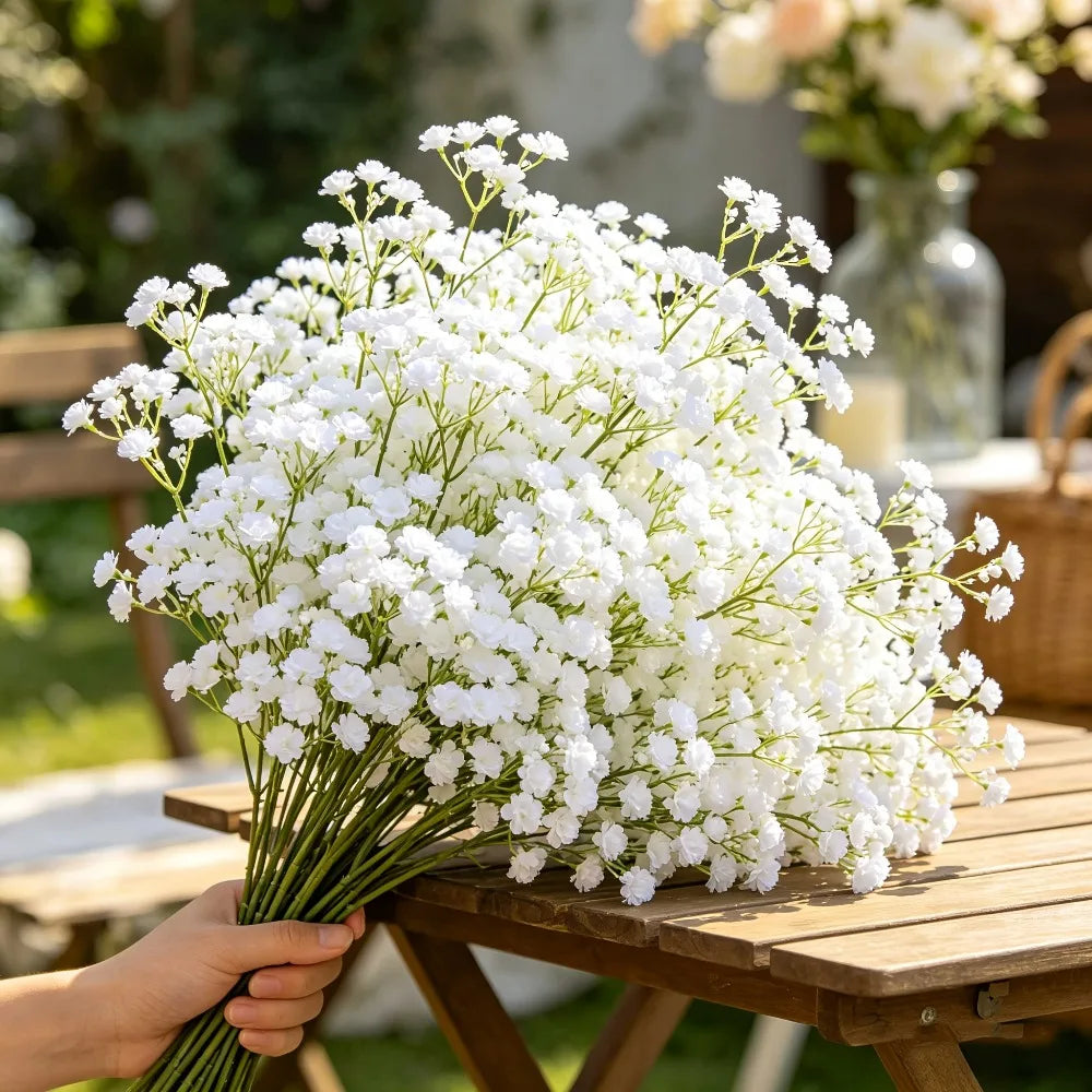 A bouquet of artificial white baby's breath flowers is held in a hand on a wooden table.
