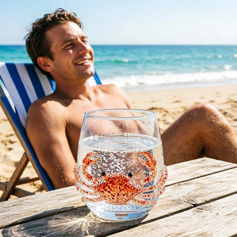 A clear glass filled with water and a preserved blue-ringed octopus decoration rests on a wooden table by the sea.