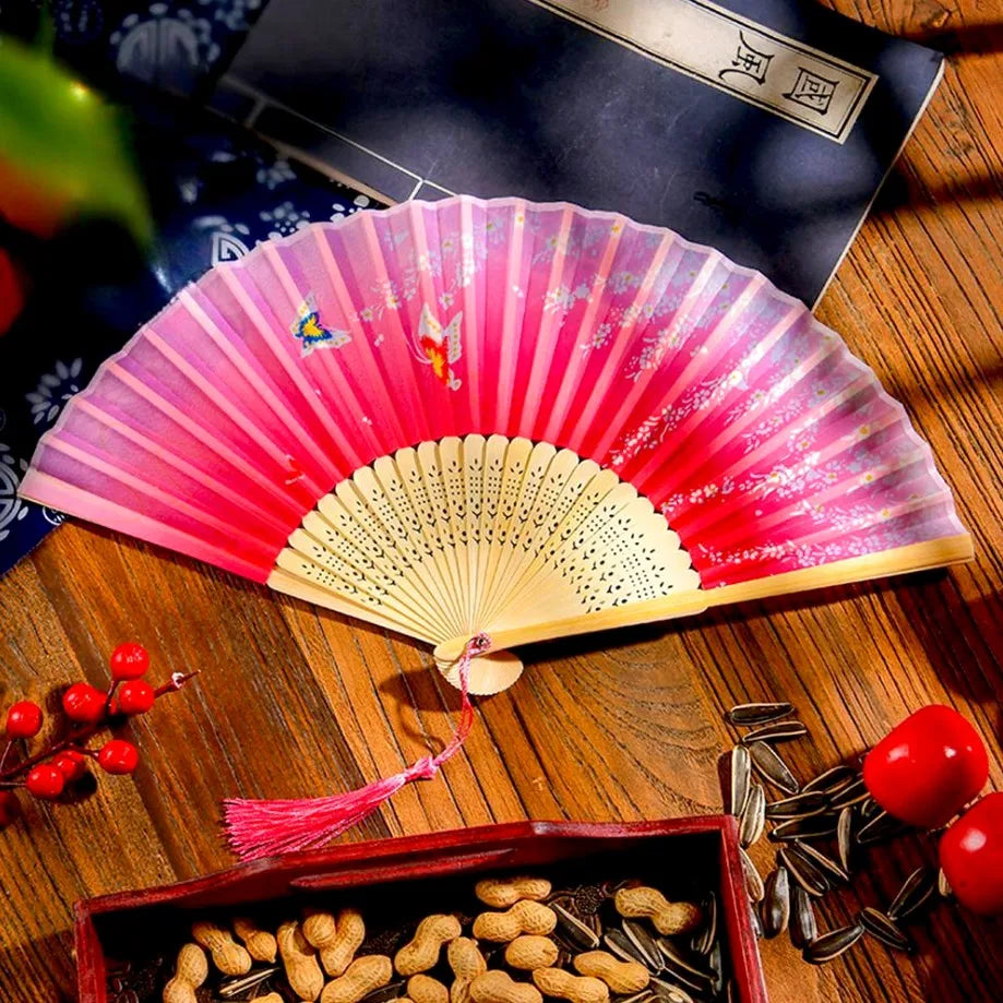 A vintage silk folding hand fan with pink and gold accents resting on a wooden surface next to dried seeds.