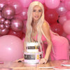 A woman in a pink dress holds a tiered cake decorated with gold accents and pearl bow toppers amidst pink balloons.