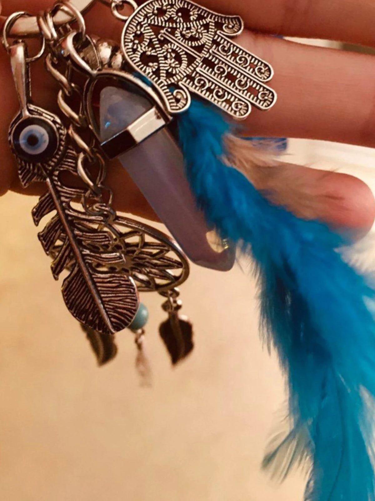 A hand holds a bohemian dream catcher keychain featuring a feather and blue tassel.