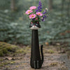 A black ceramic cat head vase holding pink and purple flowers in a natural outdoor setting.