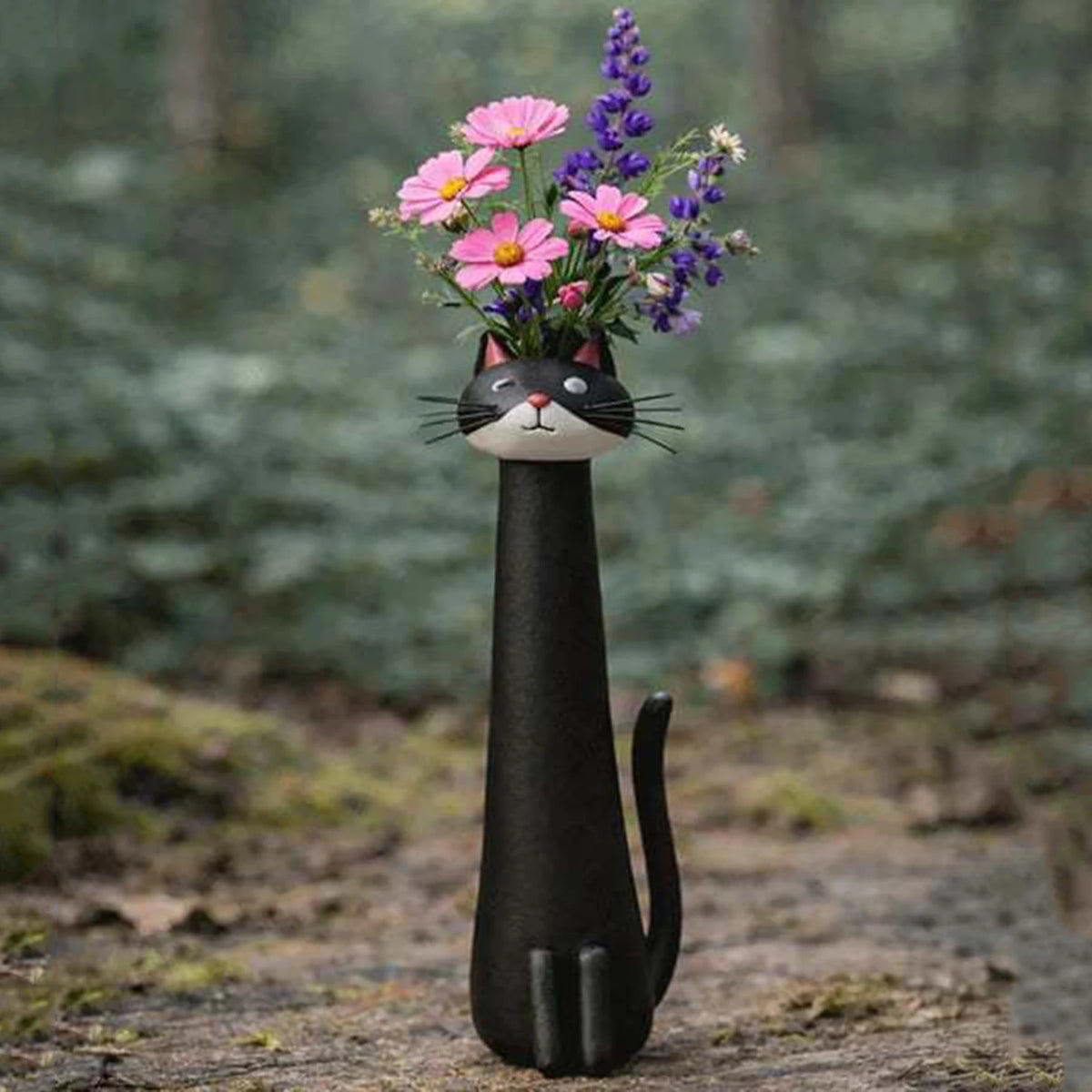 A black ceramic cat head vase holding pink and purple flowers in a natural outdoor setting.