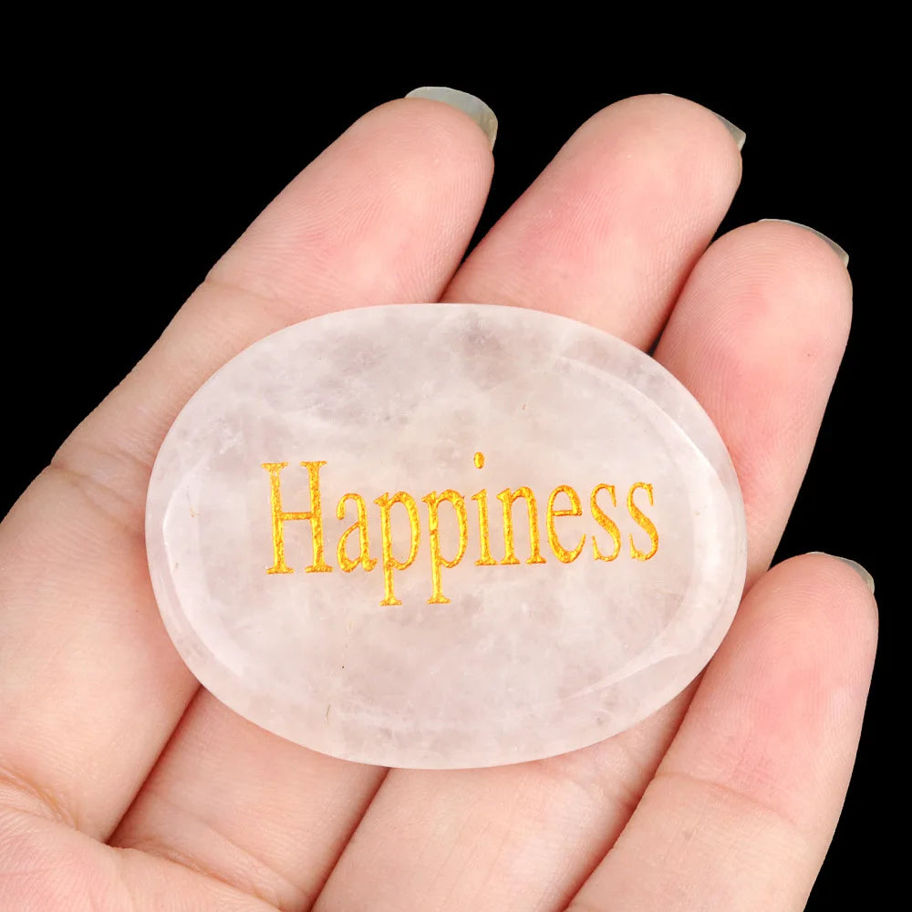A hand holds a smooth, oval-shaped pink quartz crystal with the word Happiness engraved on it.
