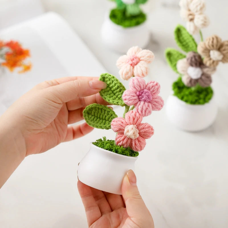 A hand holds a small white pot containing a hand-woven crochet pink flower plant.