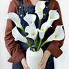 A person holds a bouquet of white calla lily artificial flowers in a white vase.
