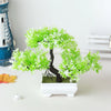 Un petit arbre bonzai artificiel avec du feuillage vert vif et un tronc sombre dans un pot rectangulaire blanc.