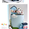 A light blue cake decorated with farm animal toppers including a white sheep, a pink pig, and a green plant.