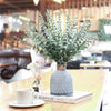 A vase filled with artificial eucalyptus leaves sits on a wooden table next to a white cup and a book.