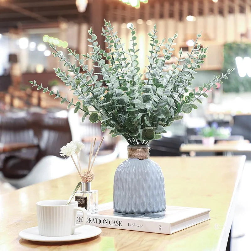 A vase filled with artificial eucalyptus leaves sits on a wooden table next to a white cup and a book.