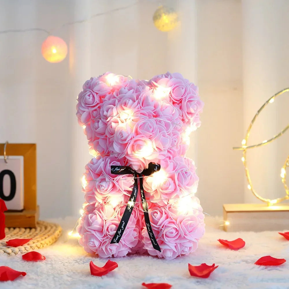A fluffy pink artificial rose teddy bear with glowing lights resting on a white surface surrounded by red rose petals.