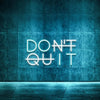 A glowing neon sign spelling DON'T QUIT is mounted on a textured blue wall with a reflective floor.
