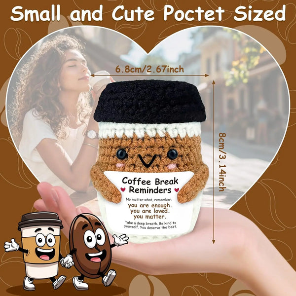 A small, handwoven knitted coffee cup cozy with a smiling doll decoration is shown next to a hand.