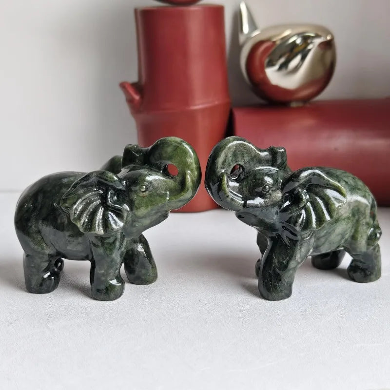 A pair of carved jade elephant statues resting on a white surface next to a red vase and silver object.