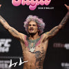 A muscular man with pink hair and tattoos stands with his arms outstretched in front of a dark background.