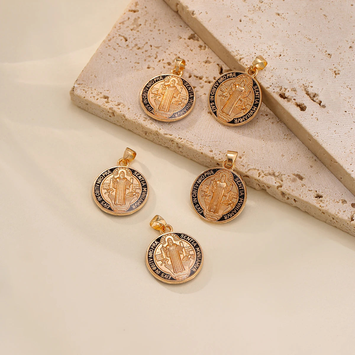 A set of five gold-toned double-sided St. Benedict medals displayed on a light stone surface.