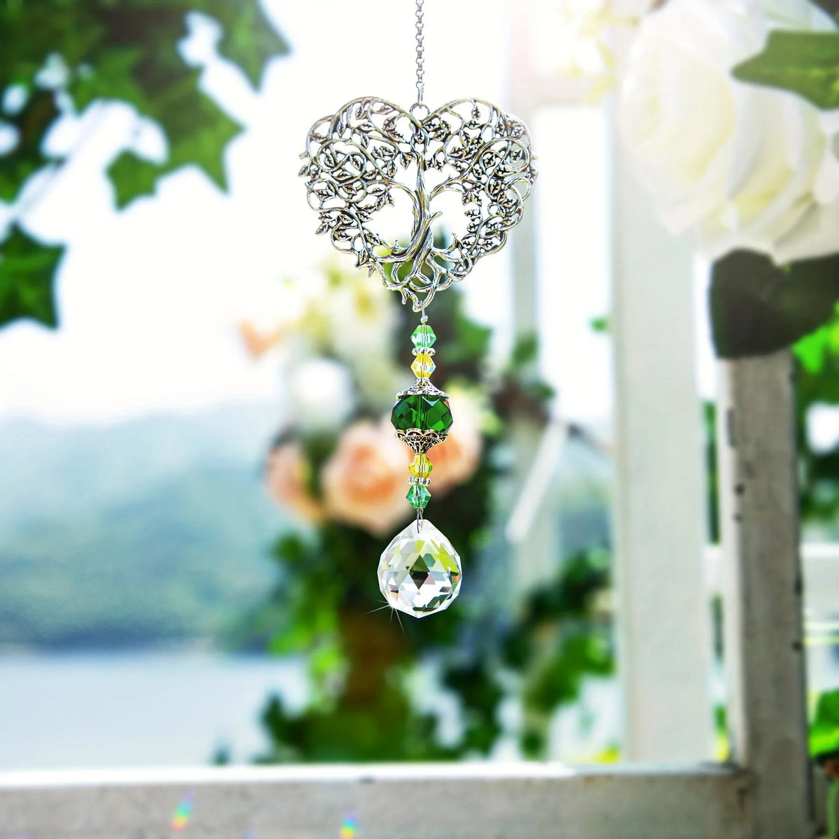 Silver crystal tree of life pendant with a green gemstone drop hanging in front of a blurred outdoor background