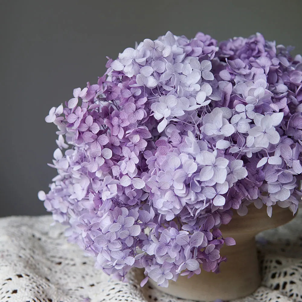 A large bouquet of purple dried hydrangea flowers arranged in a neutral vase on a textured fabric.