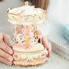 A delicate pink and white resin carousel music box with horses and floral accents being held by a person.