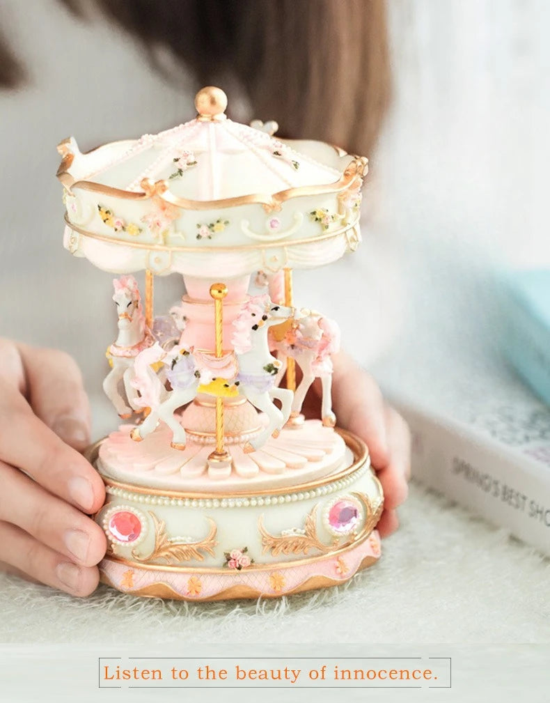 A delicate pink and white resin carousel music box with horses and floral accents being held by a person.