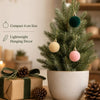 A potted green fir tree decorated with five lightweight velvet flocked ornaments in shades of green, beige, and pink.