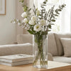 A clear glass cylinder vase filled with white roses and lavender sprigs on a wooden table.