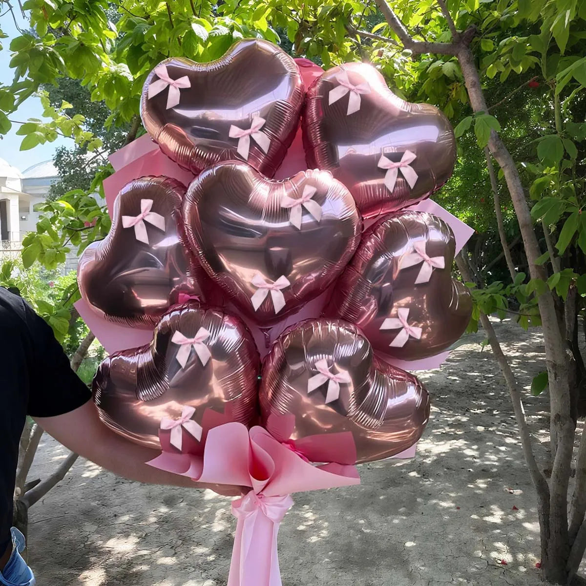 A bouquet of pink heart-shaped foil balloons tied with ribbons, held by a person outdoors.