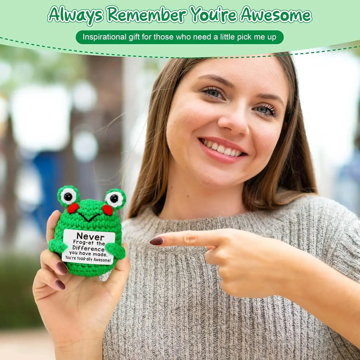 A woman holds an inspirational crochet frog ornament, pointing to it, against a blurred outdoor background.