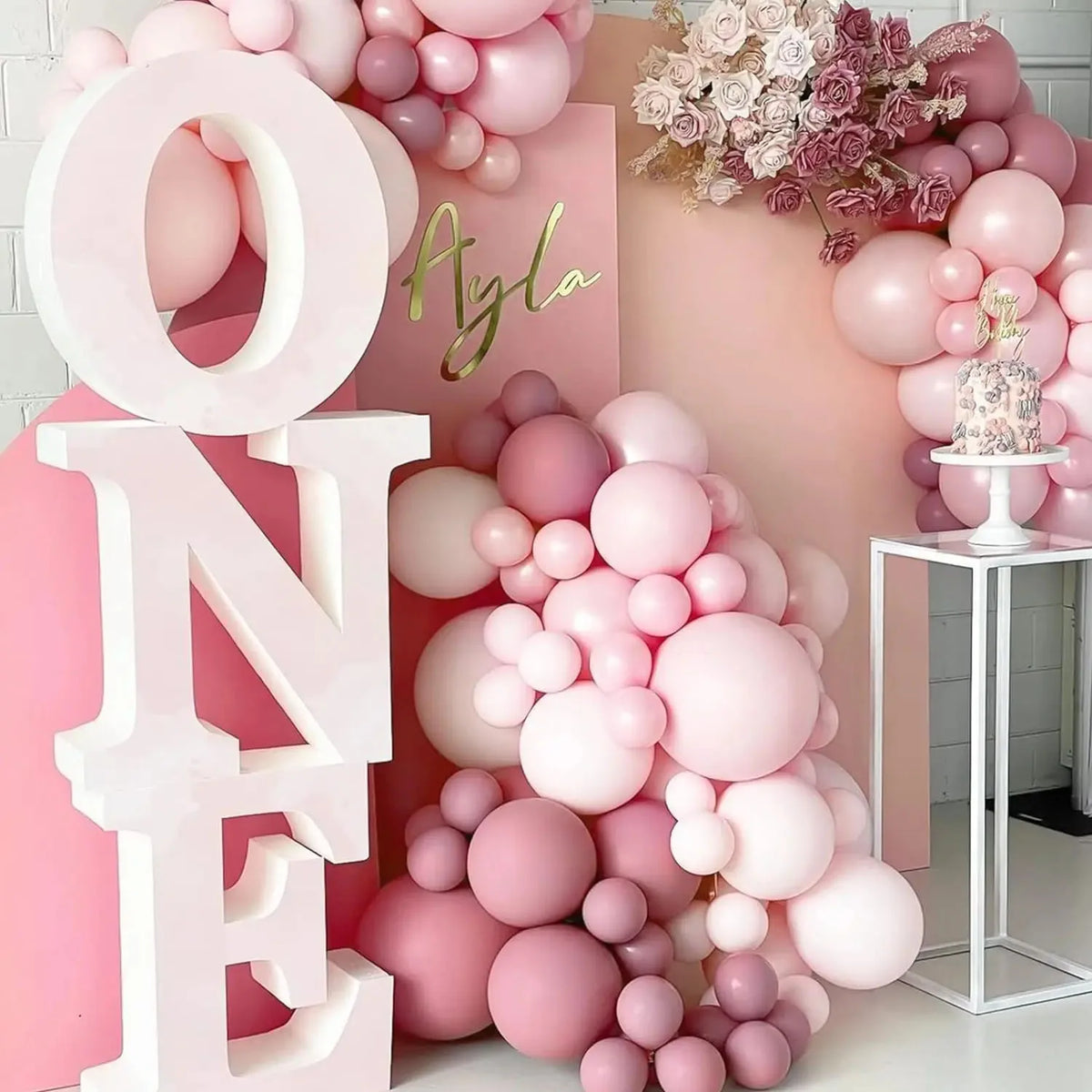 A large pink balloon letter display spelling ONE and AYLA with clusters of pastel pink balloons in a soft pink setting