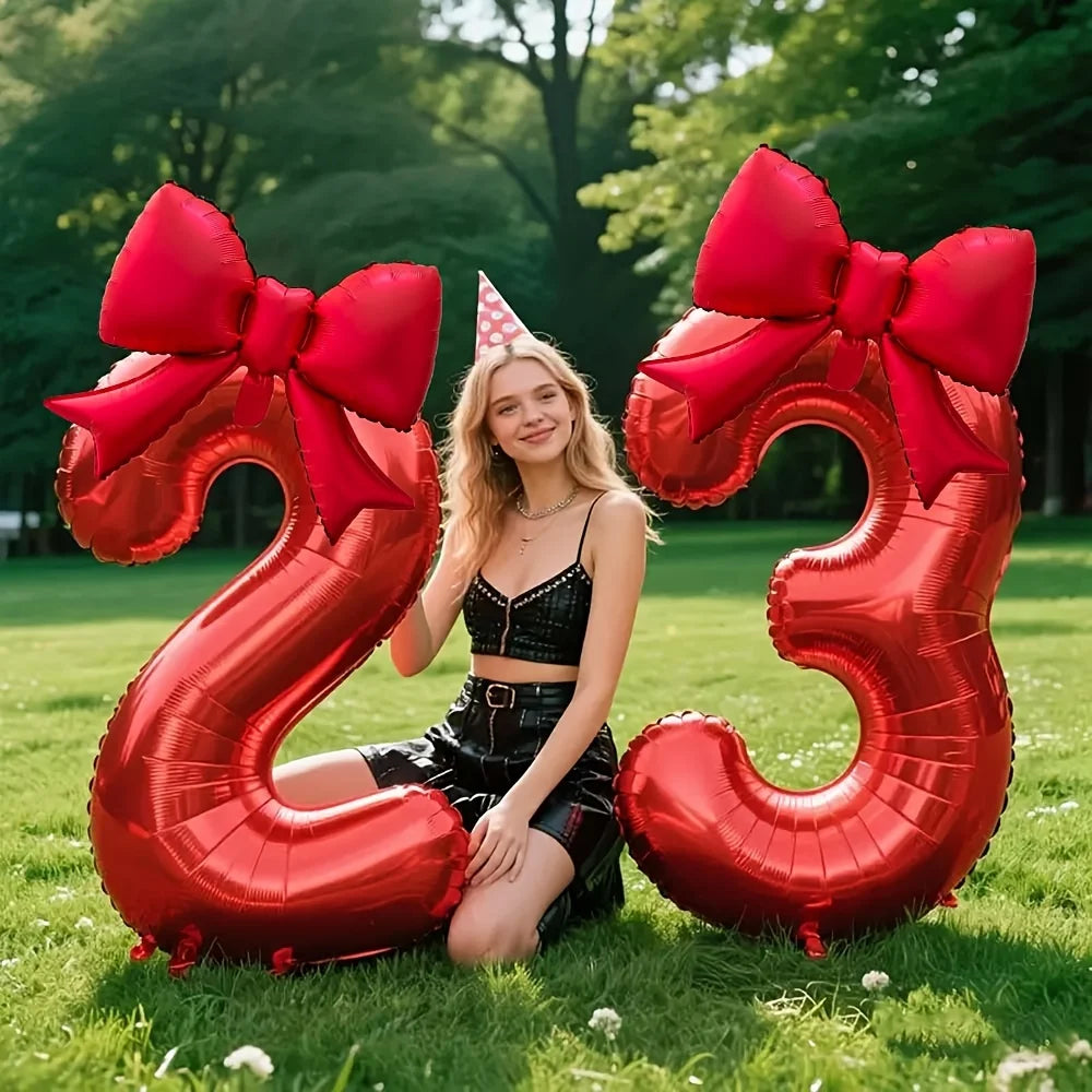 A young woman sits in front of large red foil number balloons shaped like 23, tied with matching red bows, outdoors.
