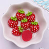 A small white dish holds several bright red strawberry resin flatback charms with green tops for DIY crafts.