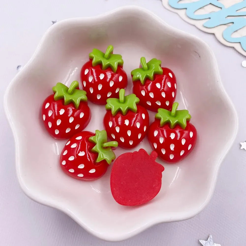A small white dish holds several bright red strawberry resin flatback charms with green tops for DIY crafts.