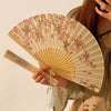 A hand holds an elegant floral bird print bamboo folding fan with light wood and pink accents.